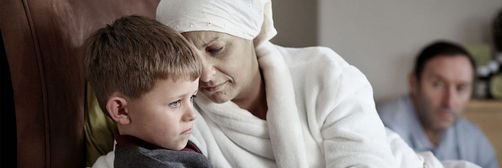 Ill lady iwith white scarf around her head in white bathrobe comforting her young son while husband looks on.