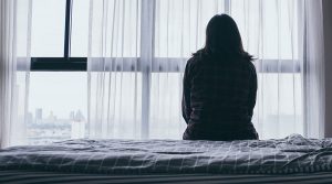 Lady sitting on bed with her back to the viewer looking out of her high rise flat windows