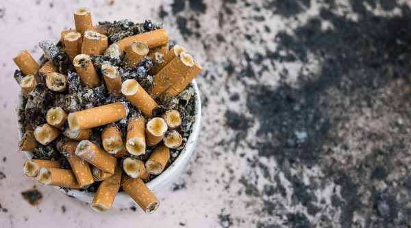 Cup filled with many cigarette ends that is sitting on the ground with cigarette ends most visible to the viewer