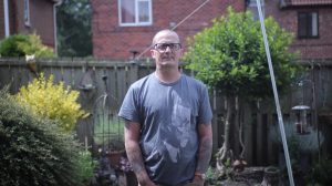 Man wearing glasses with tattoos on arms standing in garden