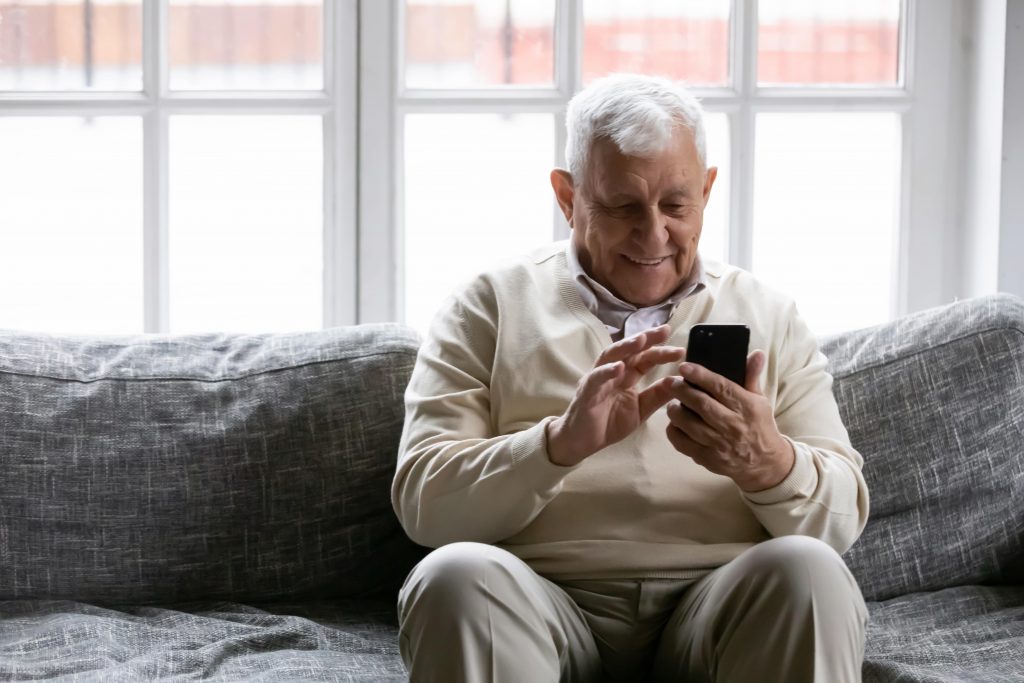 Elderly gentleman sitting on a grey sofa smiling at his smartphone will playing with it.